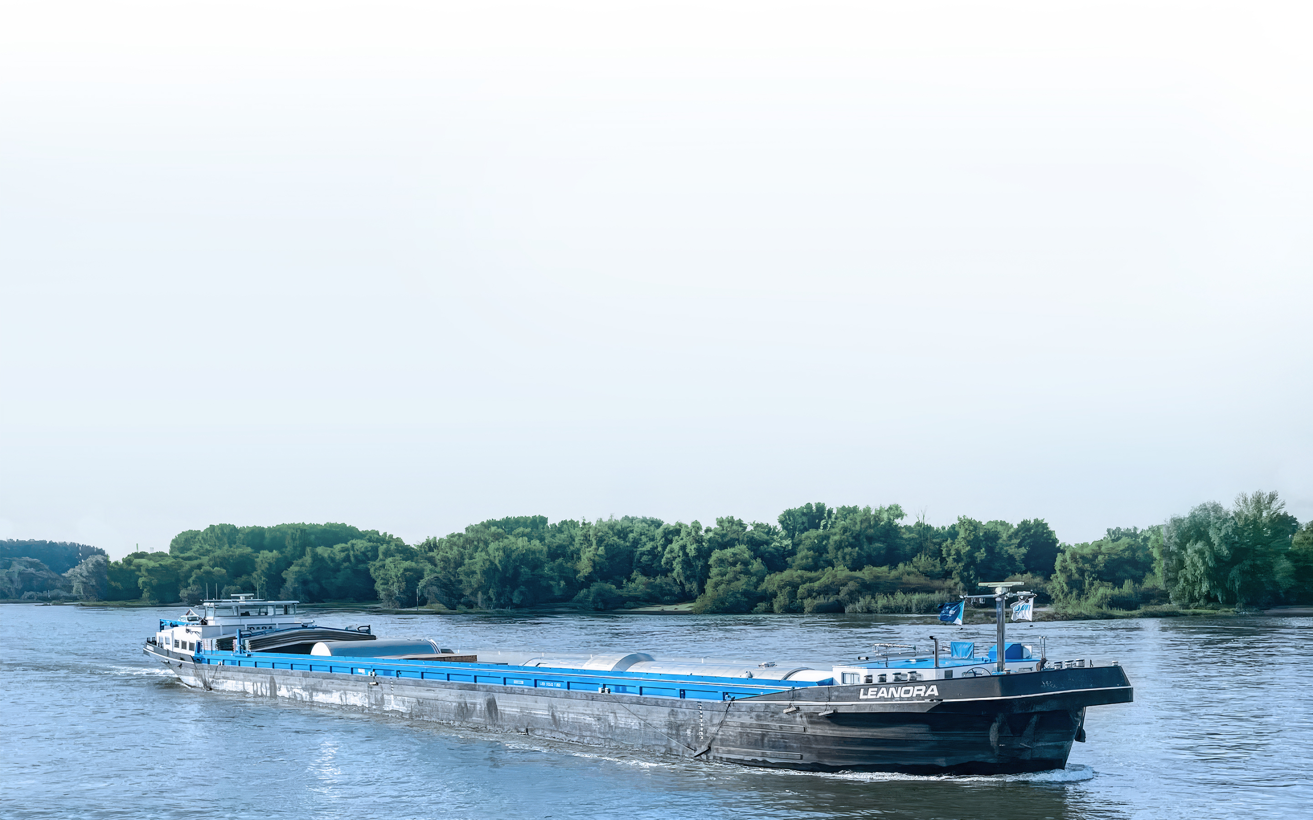 A long, black and blue barge named LEANORA navigating a wide river, surrounded by dense, green forested banks under a clear sky
